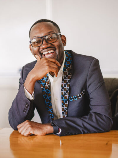 A portrait of a man with large rimmed glasses seated at a table with a big smile