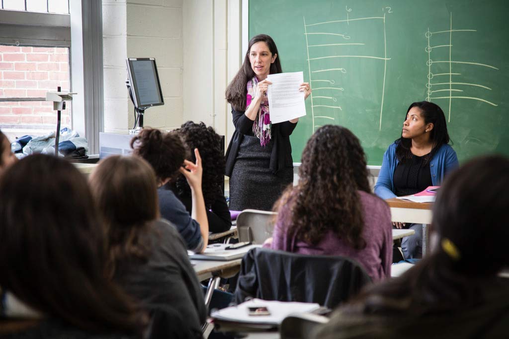 Female standing in front of chalkboard holding a piece of paper up and talking