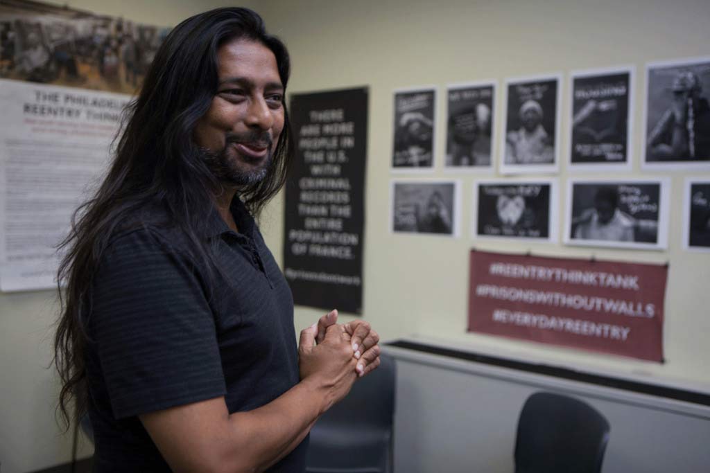 man smiling with hands clasped in front of wall of photos