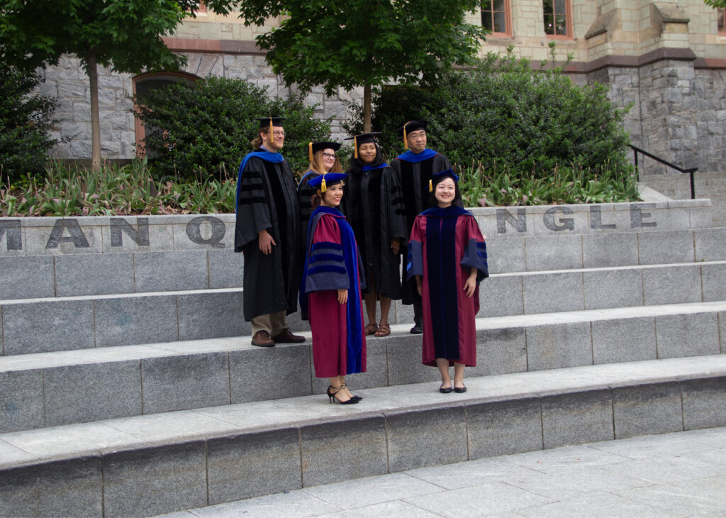Six PhD graduates standing on steps in graduation robes