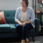 Woman in light blue blazer and white blouse sits on a turqouise couch with her arms folded and legs crossed
