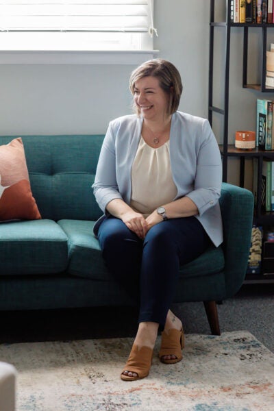 Woman in light blue blazer and white blouse sits on a turqouise couch with her arms folded and legs crossed