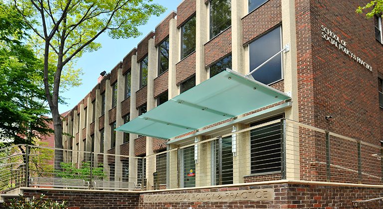 Modern brick academic building with glass canopy entrance, concrete columns, and metal railings surrounded by trees.