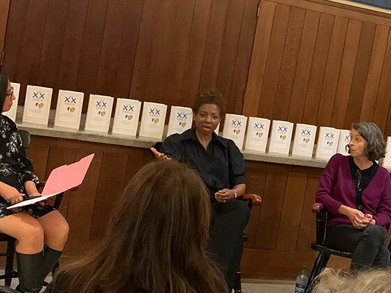 Three women in chairs converse before an audience, with a row of books on a shelf behind them