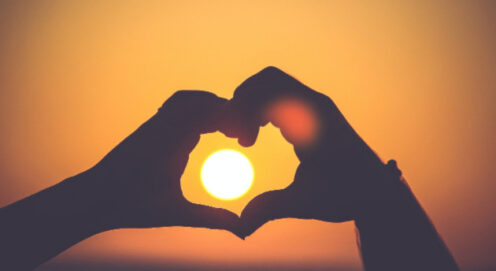 Two hands form the shape of a heart around a setting sun