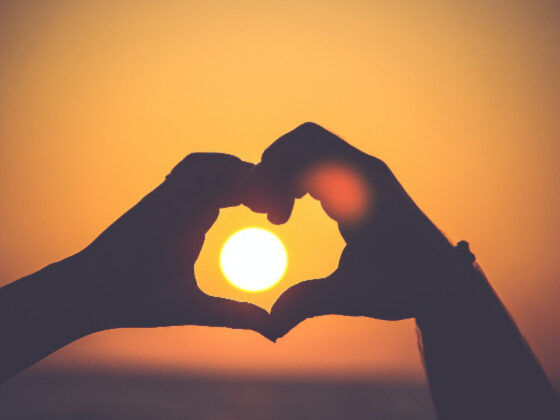 Two hands form the shape of a heart around a setting sun