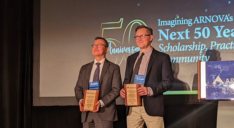 Drs. Chao Guo and Gregory Saxton stand onstage holding award plaques.