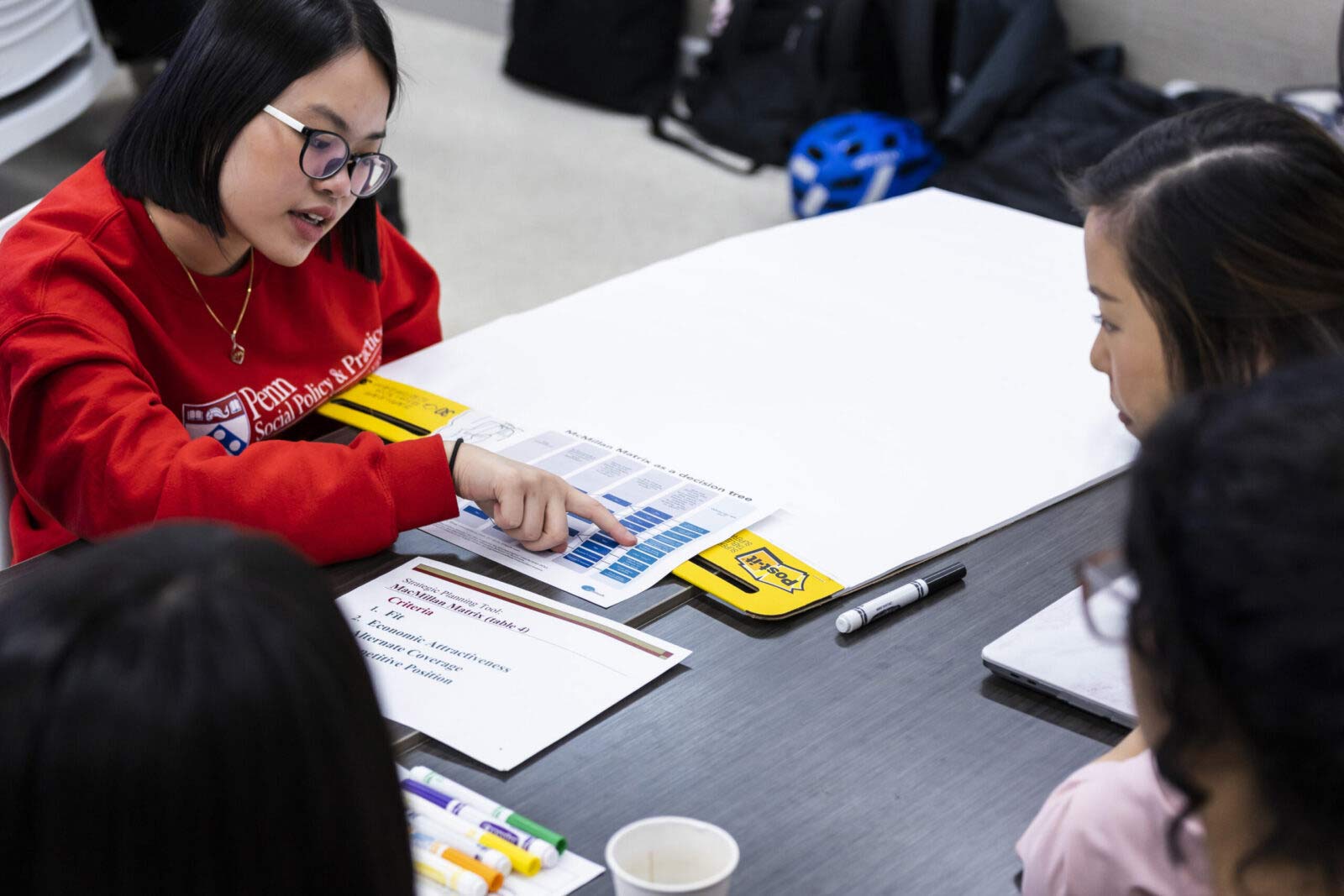 Two students sit at a table. One points at a document that sits between them.