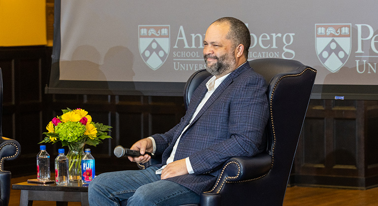 Ben Jealous sits onstage smiling at the audience. Behind him on a blue screen appears the logo of the Annenberg School.