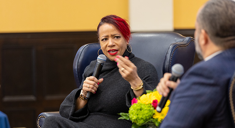 Nikole Hannah-Jones sits onstage speaking into a microphone and gesturing with one hand. Ben Jealous is shown from behind, sitting across from her.