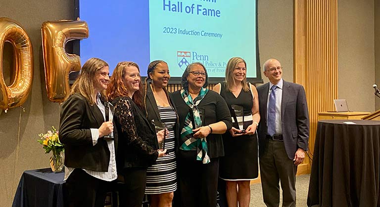 Five people pose in a row. Behind them, a screen says "Hall of Fame."