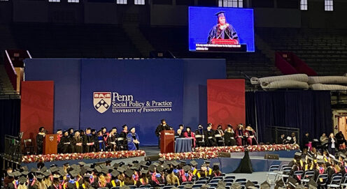 Amber Hikes speaks at a podium and projected onscreen before an audience of graduates at the Palestra, under the Penn Social Policy & Practice logo