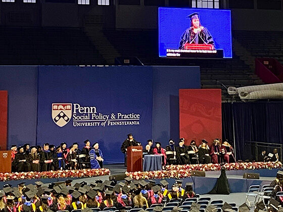 Amber Hikes speaks at a podium and projected onscreen before an audience of graduates at the Palestra, under the Penn Social Policy & Practice logo
