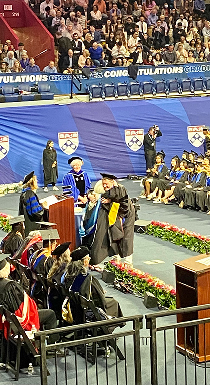 Onstage at the Palestra, two people hug before the faculty. Graduating students sit in rows and the audience sits in bleachers.