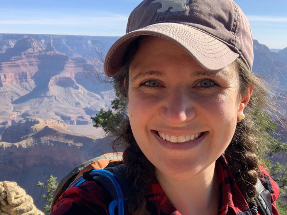 Headshot of Millan AbiNader at the Grand Canyon. Millan is wearing a hat, backpack, and red plaid shirt.