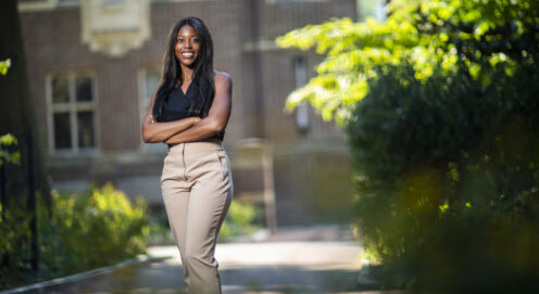 Gianni Morsell posing on campus. Gianni is standing in front of a brick building with a green plant next to her. Her arms are crossed and she is wearing a black shirt and khaki pants.