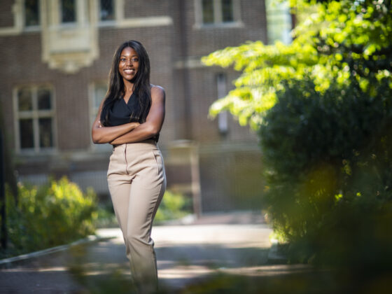 Gianni Morsell posing on campus. Gianni is standing in front of a brick building with a green plant next to her. Her arms are crossed and she is wearing a black shirt and khaki pants.