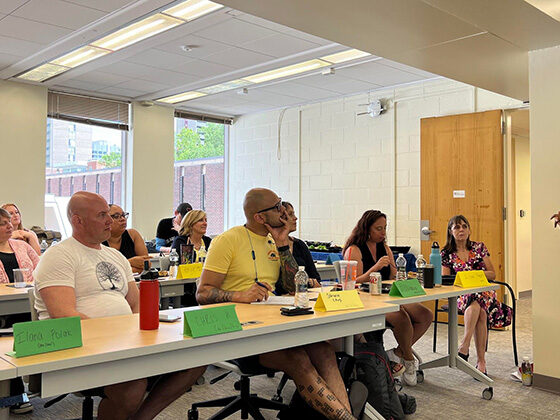 Adult students sit in rows at tables with colorful nametags, listening to a speaker