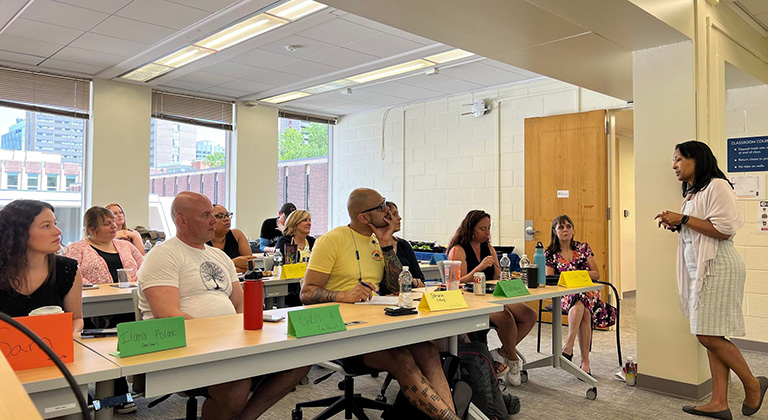 Adult students sit in rows at tables with colorful nametags, listening to a speaker