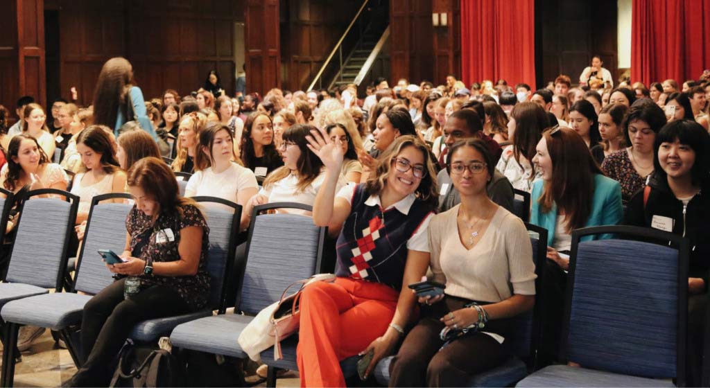 Two SP2 students sitting in the front row of a packed Hall of Flags wave at the camera.