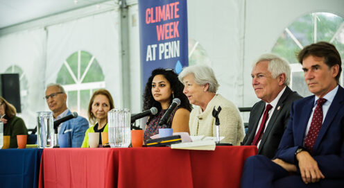 Dean Bachman speaks into a microphone, seated among panelists in a tent in front of a Climate Week at Penn banner