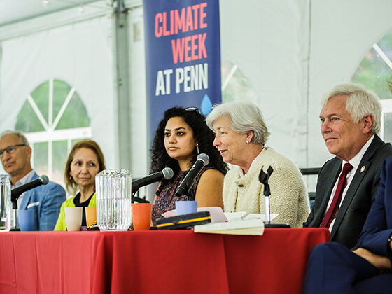 Dean Bachman speaks into a microphone, seated among panelists in a tent in front of a Climate Week at Penn banner