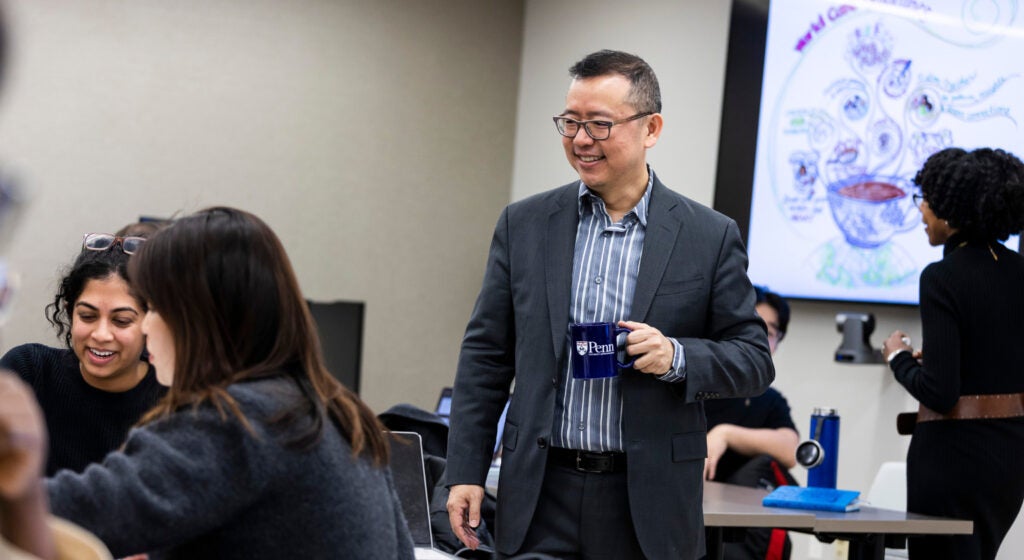 Chao Guo stands smiling and holding a Penn mug in the middle of a classroom where graduate students are seated.