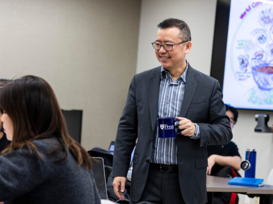 Chao Guo stands smiling and holding a Penn mug in the middle of a classroom where graduate students are seated.