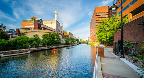 Photo of the Broad Canal in Cambridge, Massachusetts, shows a body of water with sidewalks and buildings on either side and a blue sky above