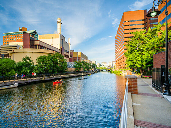 Photo of the Broad Canal in Cambridge, Massachusetts, shows a body of water with sidewalks and buildings on either side and a blue sky above