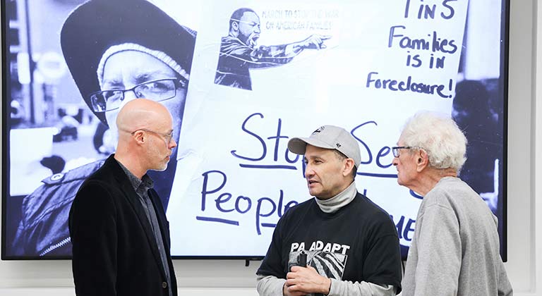 Bart Miltenberger, Harvey Finkle, and a person wearing a PA ADAPT T-shirt stand in conversation with a black-and-white photo of a disability rights activist onscreen behind them.