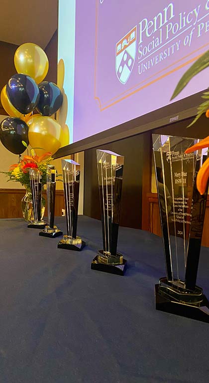 Five glass award sculptures sit in a row on a table under a screen with the SP2 logo and balloons in the background.