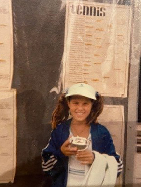 Childhood photo of Jennifer Prah standing in front of a bulletin board that says "tennis" and holding an ice cream