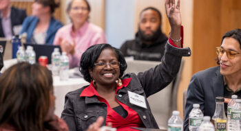 An audience member seated at a round table raises her hand, smiling