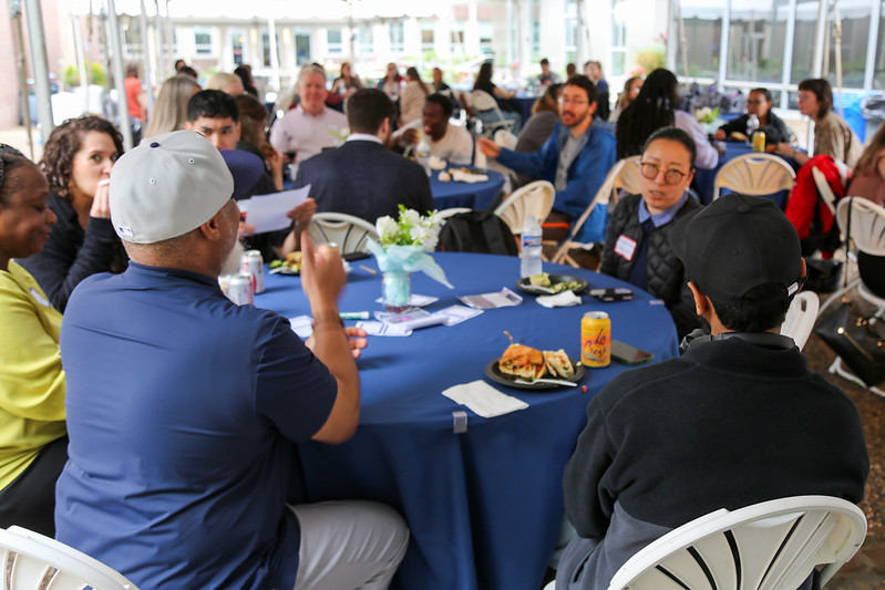 Photo of a group of students, staff and faculty in the midst of a conversation, gathered at a table with a blue tablecloth, under a tent in a courtyard.