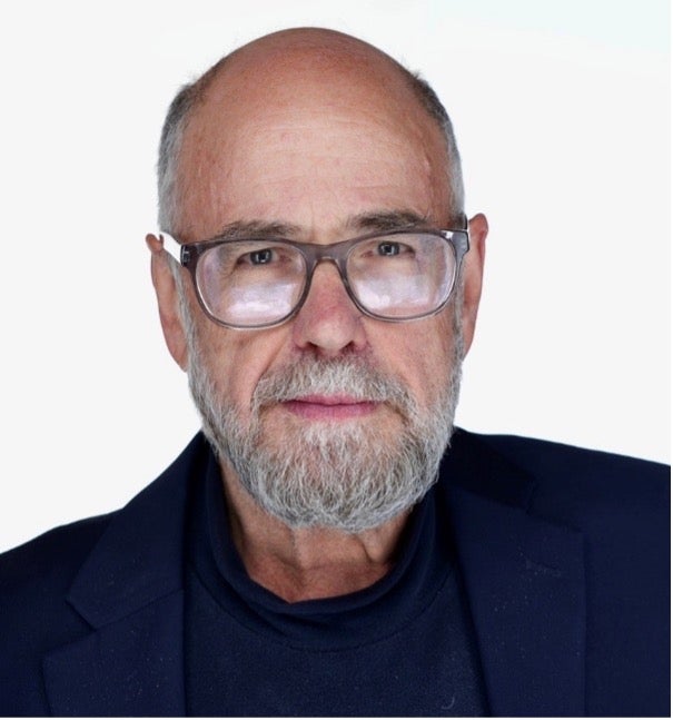 A portrait of a gray-bearded man with a square silver glasses in a navy blue shirt and suit jacket